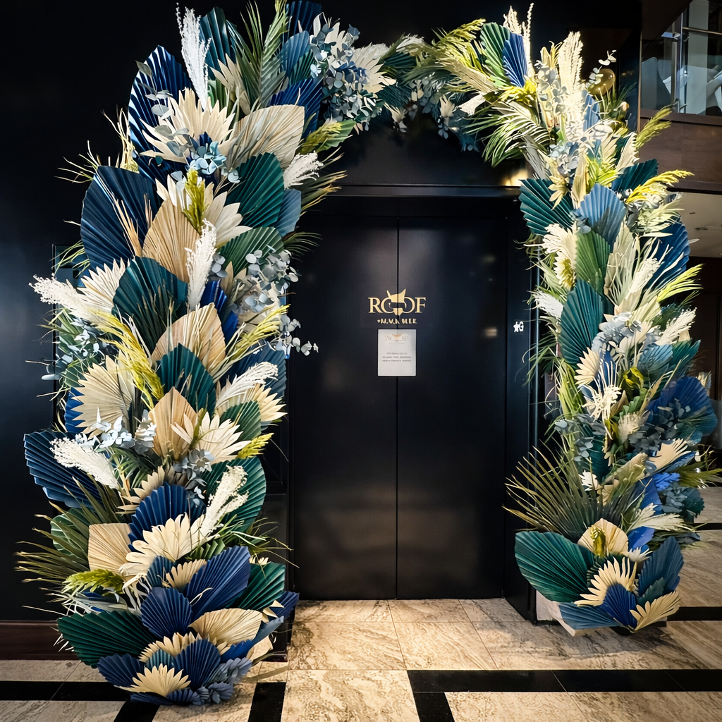 Large entrance arch installation with blue, teal, and natural dried palm fans, grasses, and foliage framing a dark doorway in an indoor venue.