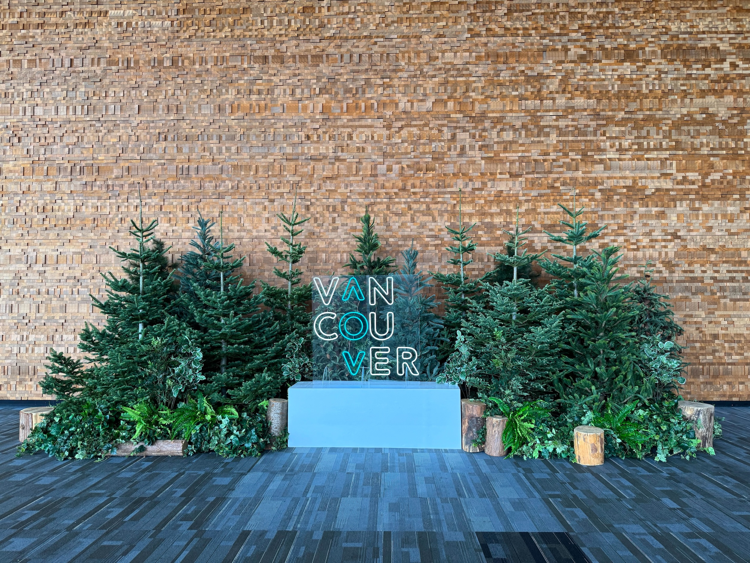 Backdrop of evergreen trees, low greenery, and wood stump accents with an illuminated Vancouver sign in front of a wood feature wall.