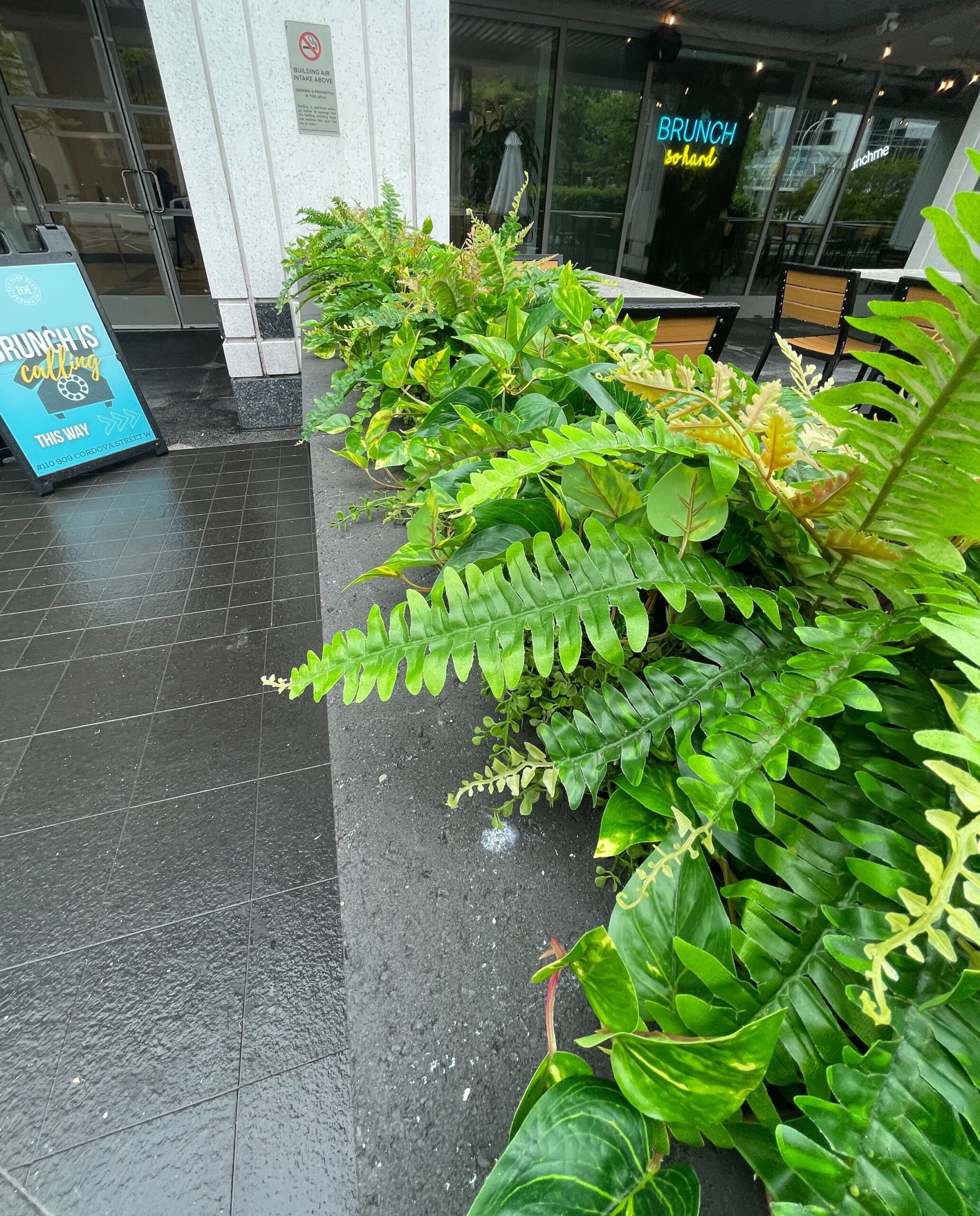 Concrete planter filled with artificial ferns, faux foliage and layered green leaves beside a commercial restaurant patio.