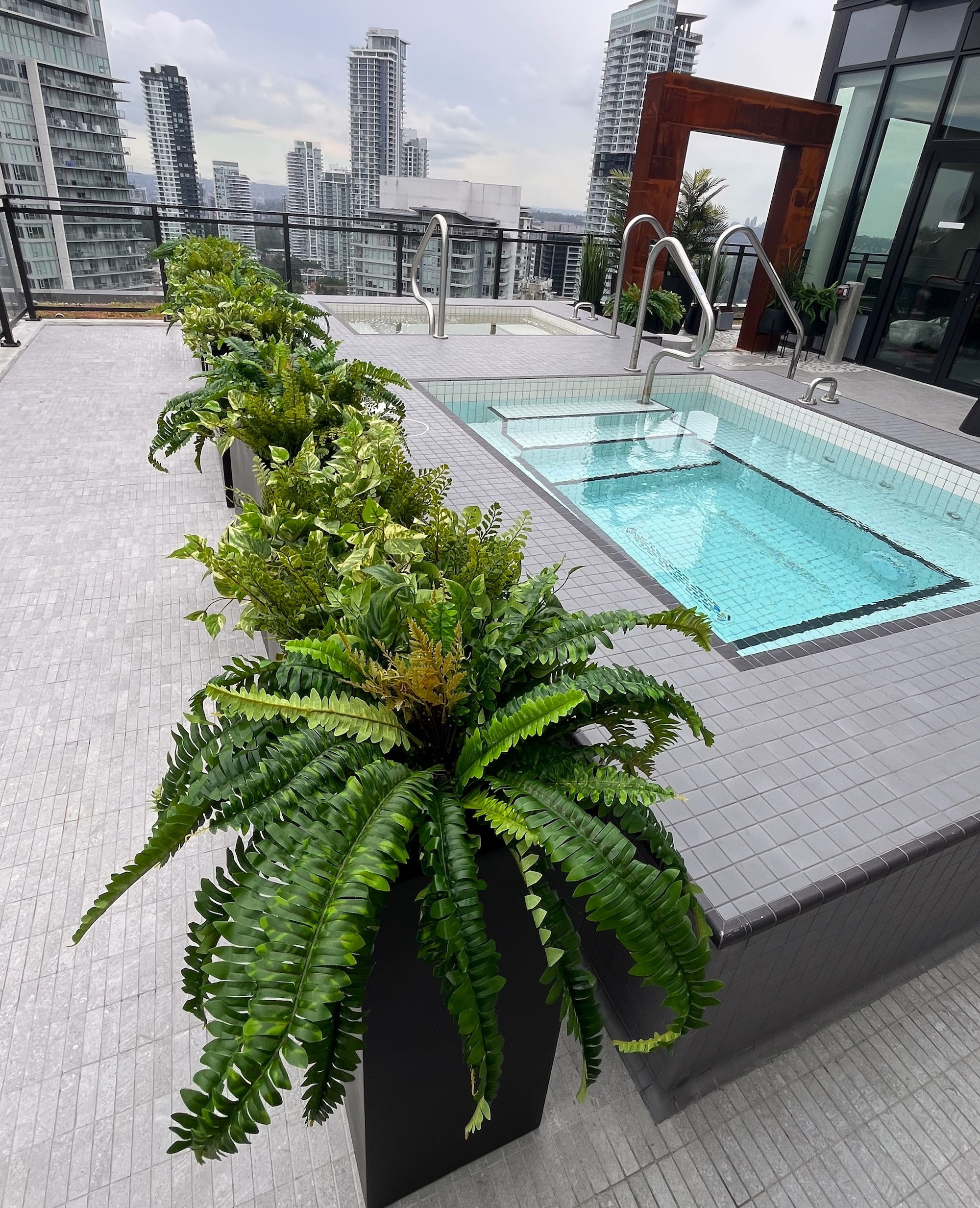Black rectangular planters filled with faux ferns and mixed artificial greenery beside a rooftop pool on a high-rise terrace.