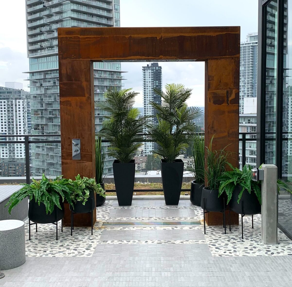 Artificial greenery in black planters arranged around a rusted steel frame on a rooftop terrace