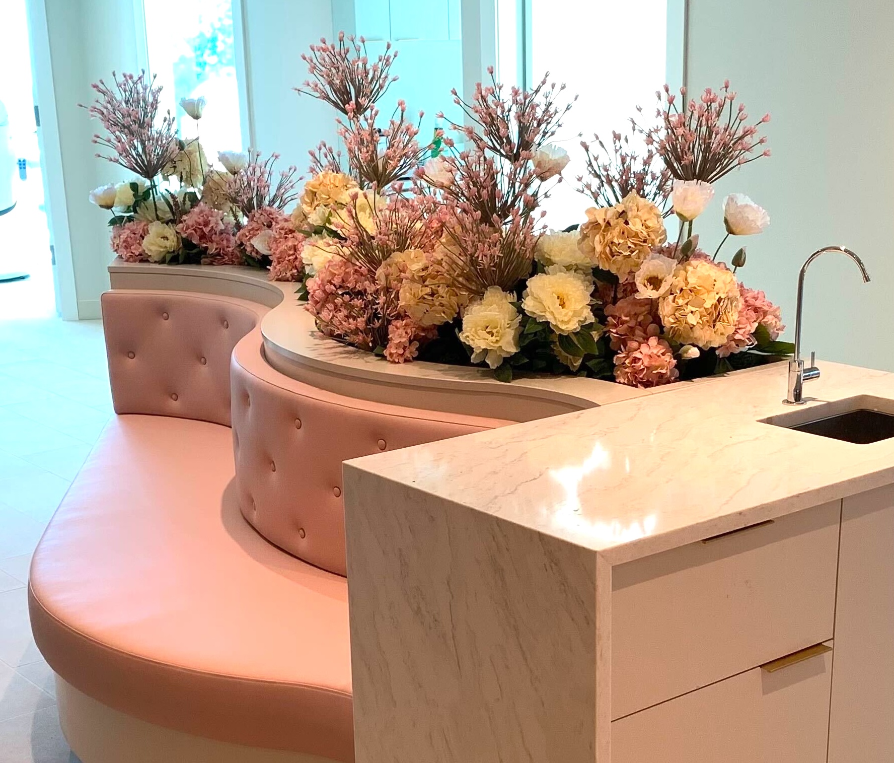 Curved built-in lobby seating with integrated faux floral planters filled with pink and cream blossoms and tall branching stems.