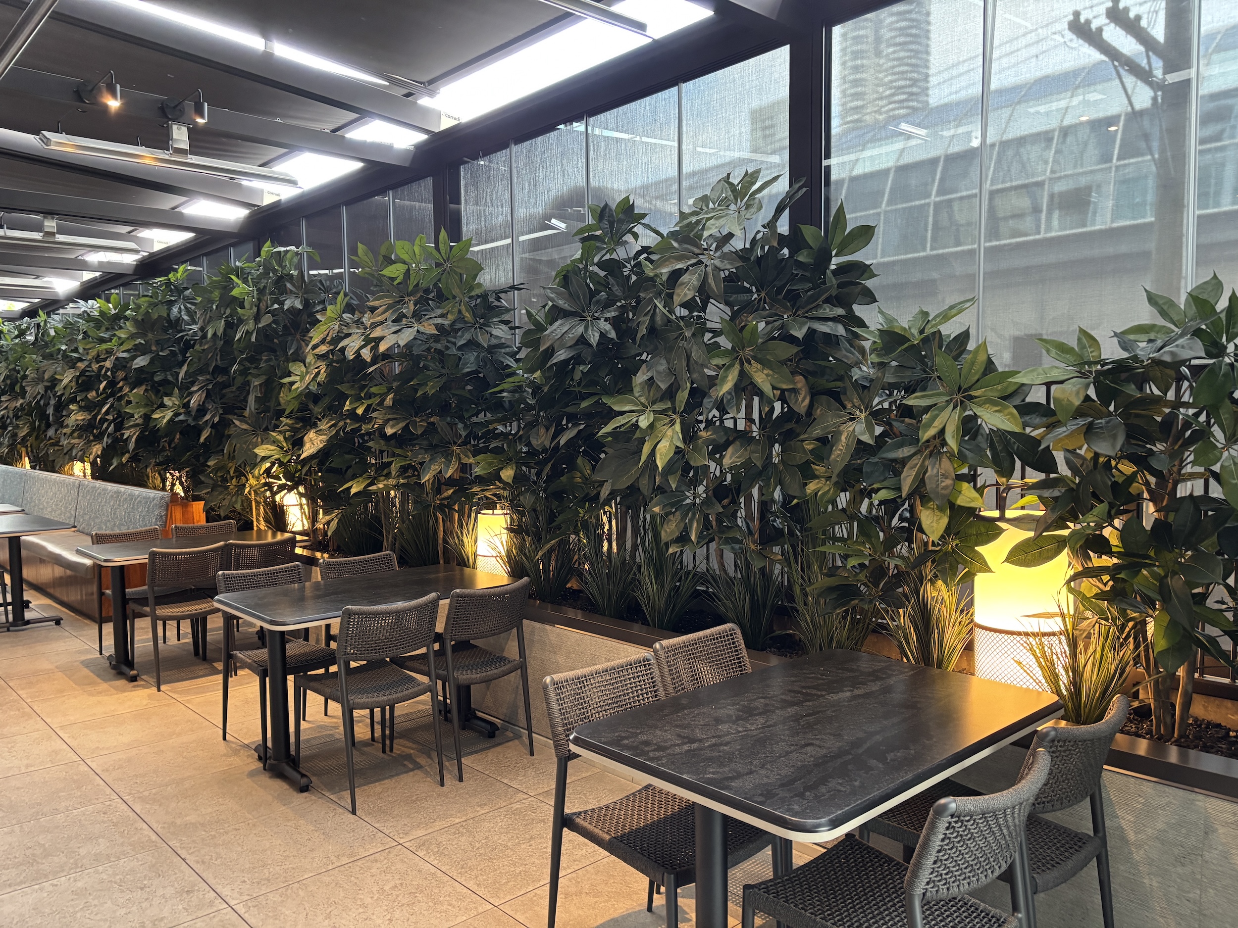 Long planter with tall faux greenery and grasses forming a screen beside restaurant patio tables and chairs.