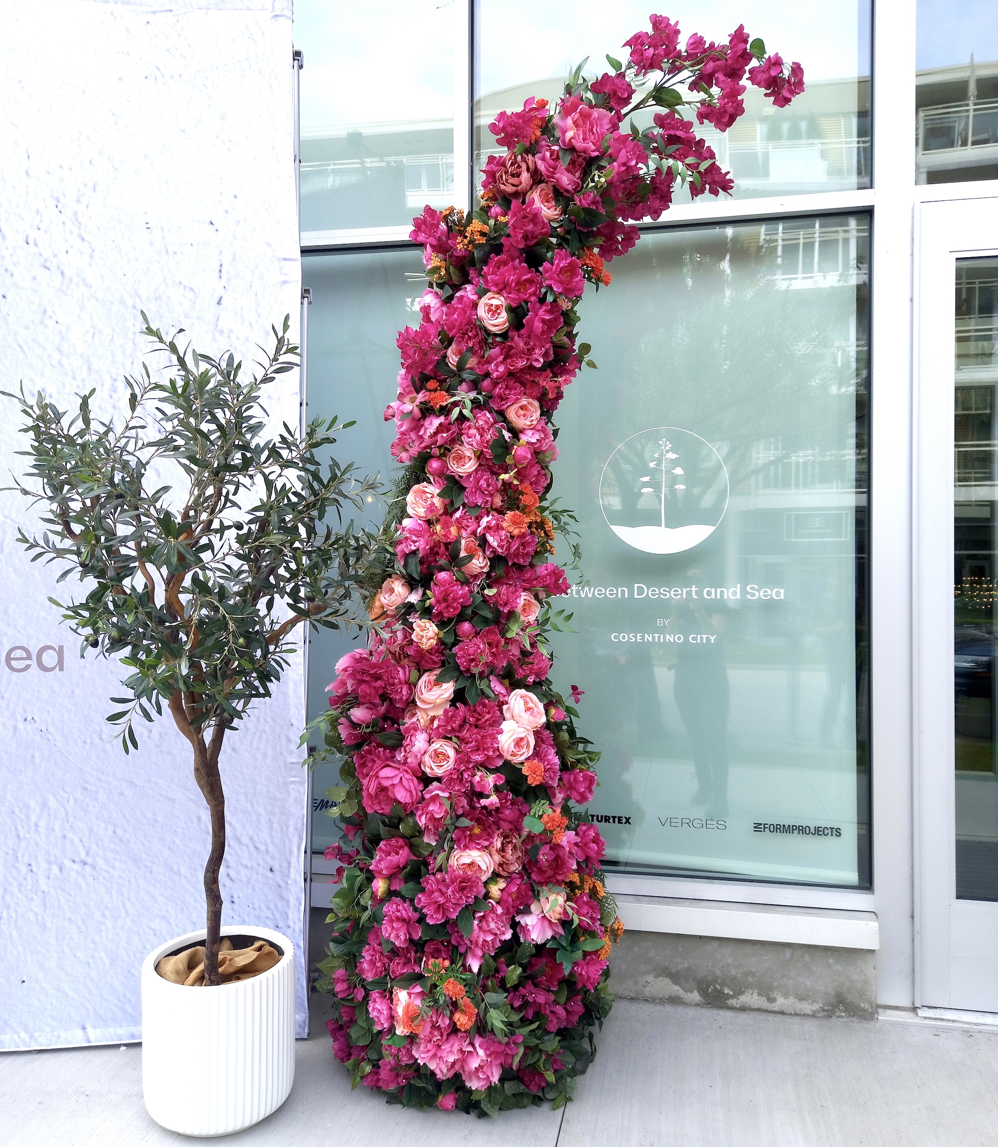 Tall floral display column covered in bright pink, blush, and coral flowers beside a potted artificial tree outside a glass-front venue.