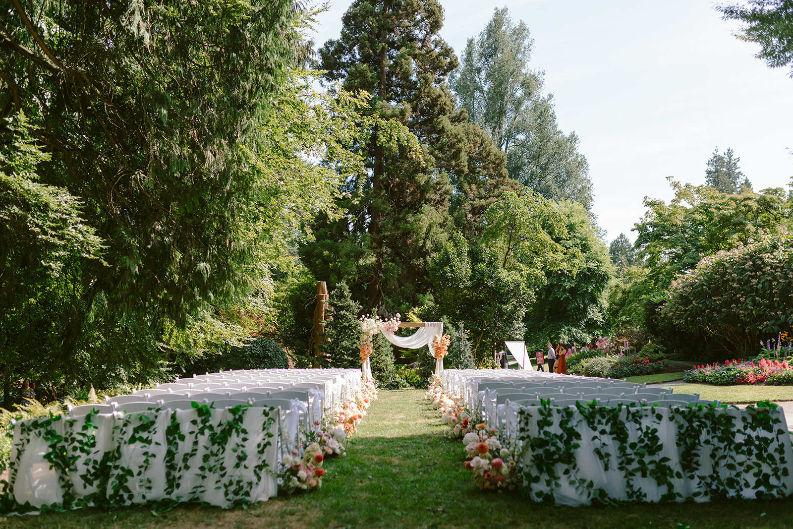 Outdoor wedding ceremony setup with white chairs, white draped aisle panels covered in green vines, pastel floral arrangements, and a floral arch on the lawn.