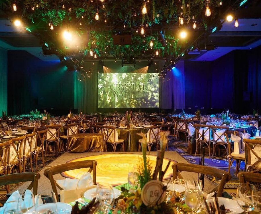 Ballroom with a hanging greenery canopy and suspended Edison bulbs above dining tables, with a stage screen at the far end of the room.