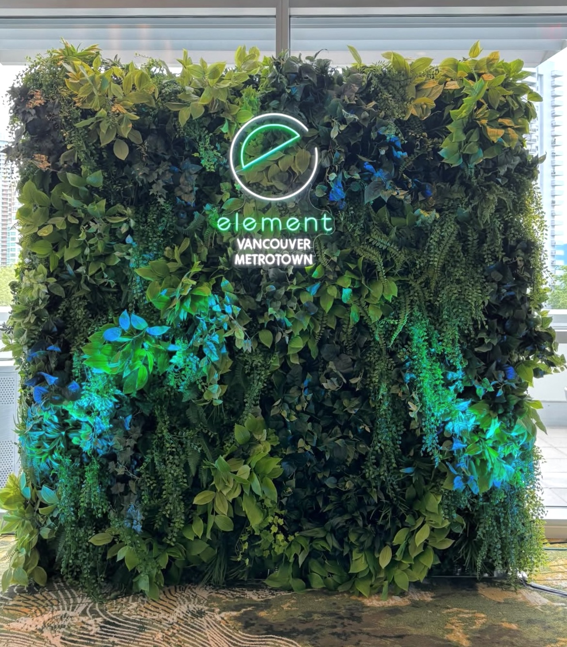 Greenery wall backdrop with mixed faux leaves and trailing foliage, featuring an illuminated sign in front of large windows.