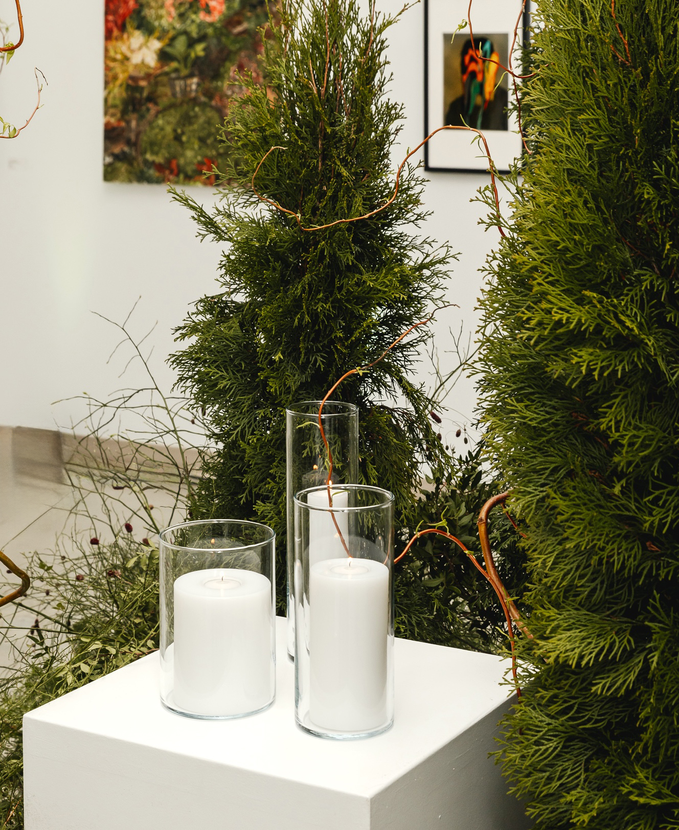 White pedestal styled with sculpted evergreen forms, airy branch accents, and layered greenery in a corporate event setting.