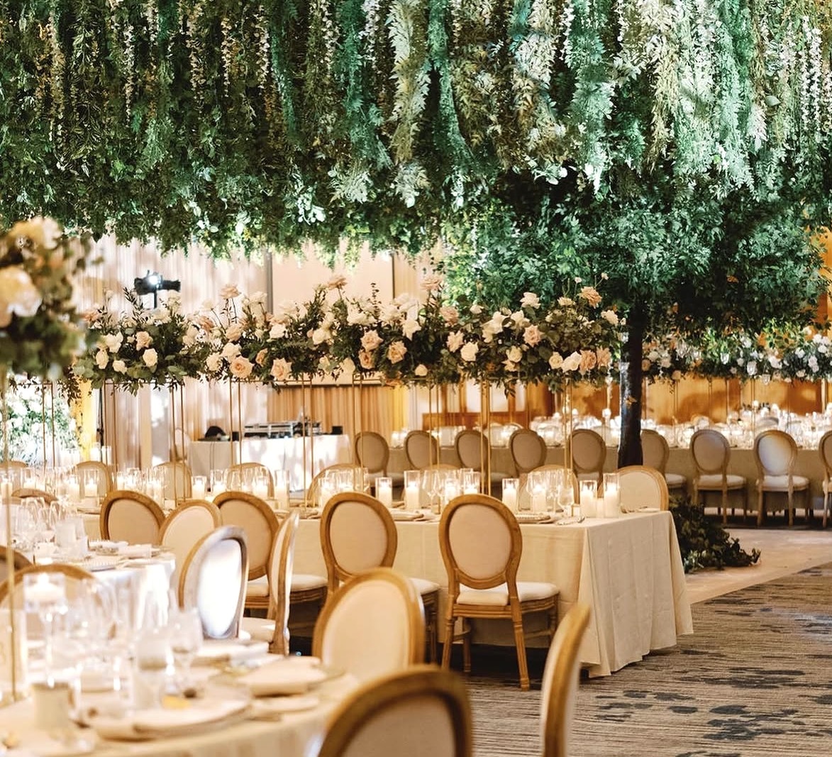 Indoor reception space with suspended green foliage canopy above dining tables, floral centrepieces on tall gold stands, and cream upholstered chairs.