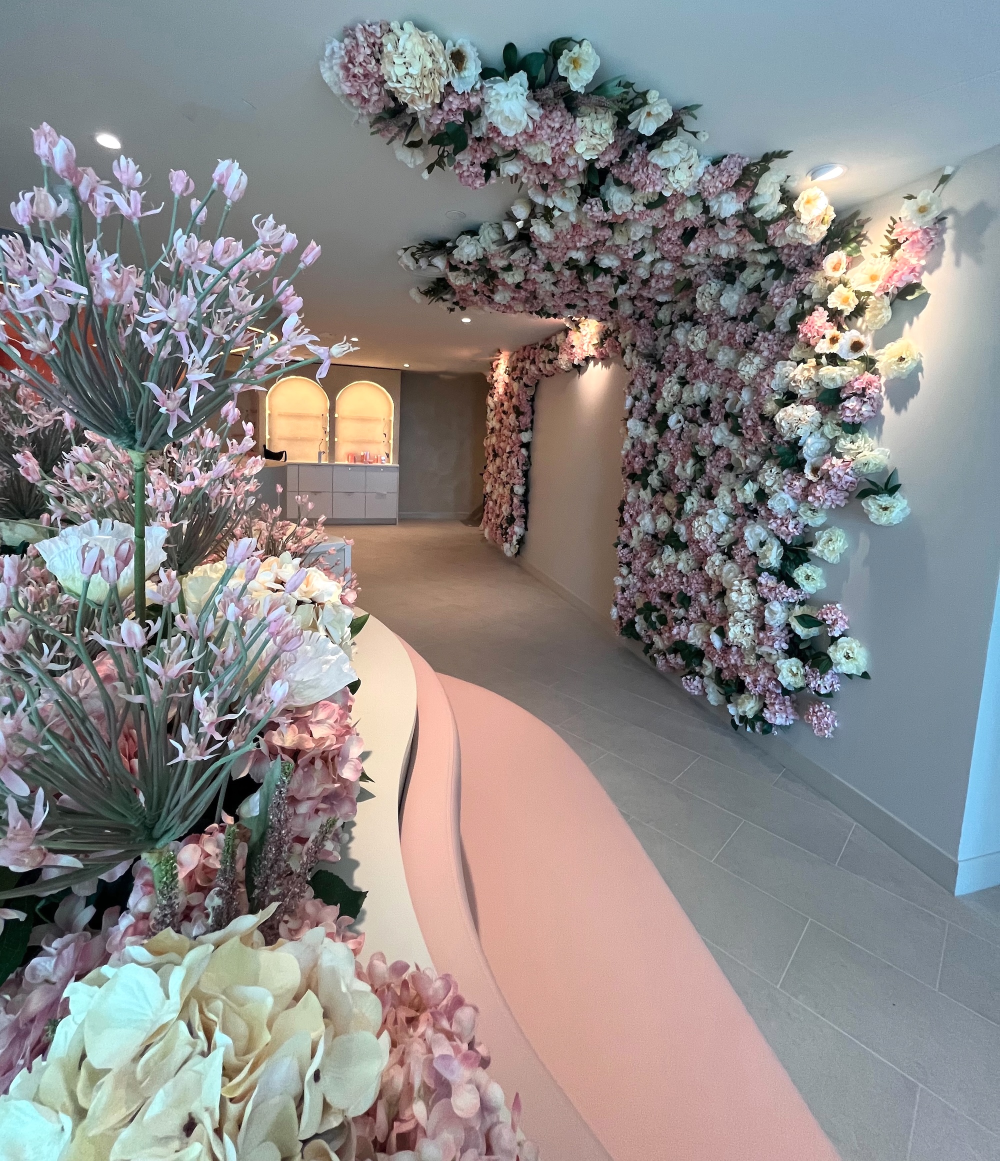 Medical office lobby with pink and white faux floral wall and ceiling installation, and blush flower arrangements along a curved reception desk.