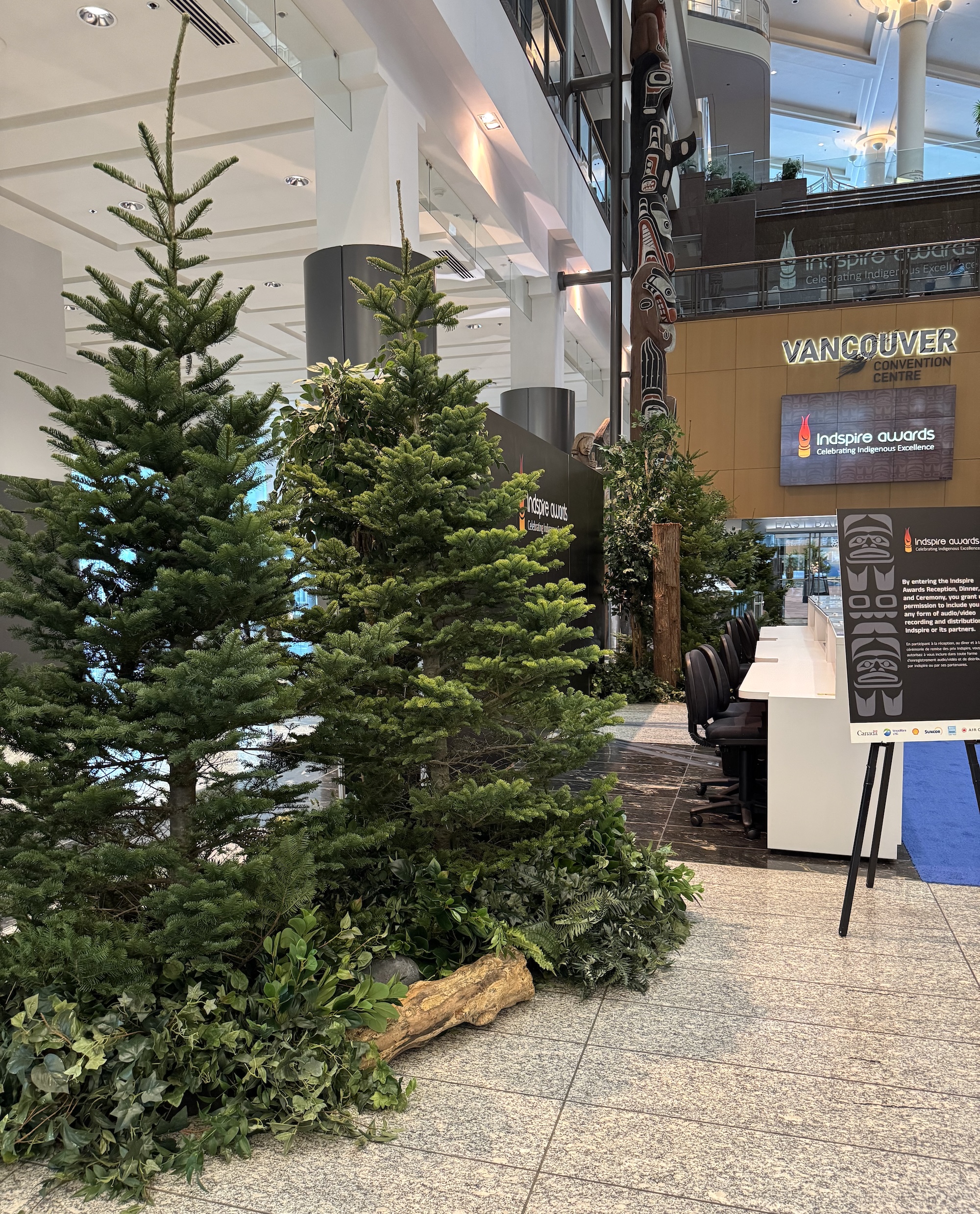 Layered evergreen trees, leafy ground greenery, ferns, and driftwood arranged in a corporate event venue beside a totem pole.