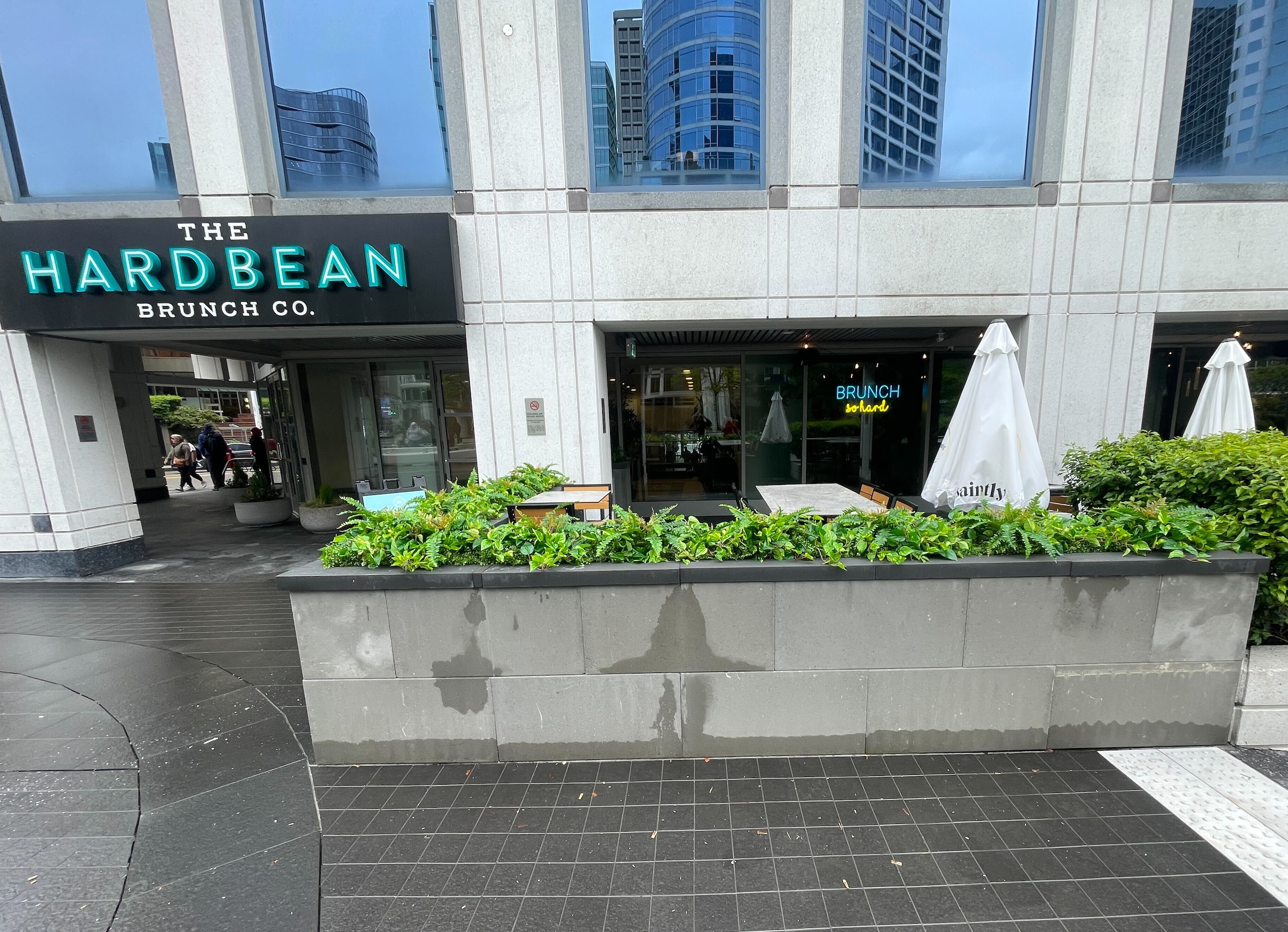 Long concrete planter boxes filled with artificial ferns and mixed faux greenery border a restaurant patio in front of a commercial building.