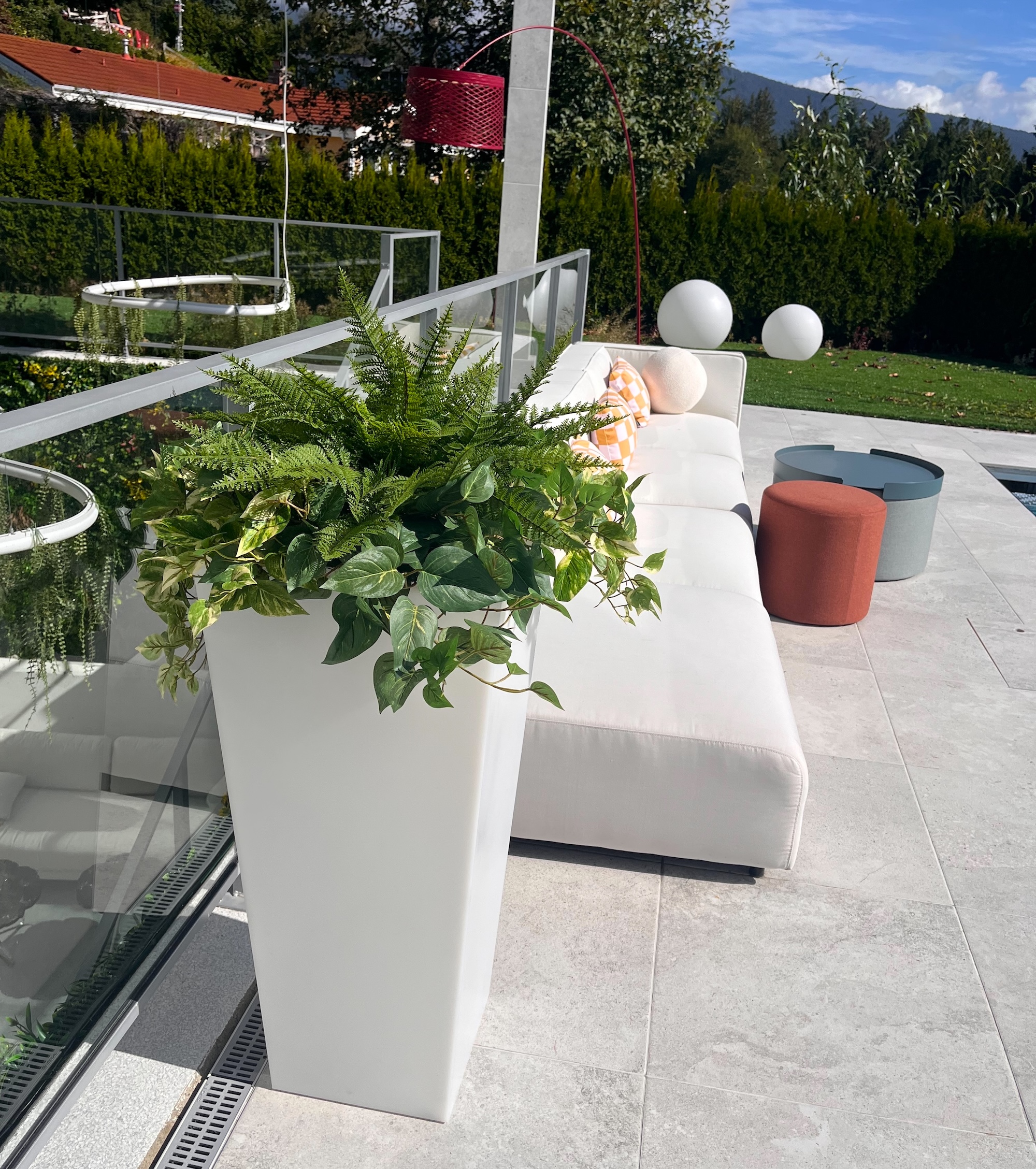 Tall white planter filled with artificial ferns and trailing faux greenery beside a white outdoor lounge sofa on a patio.