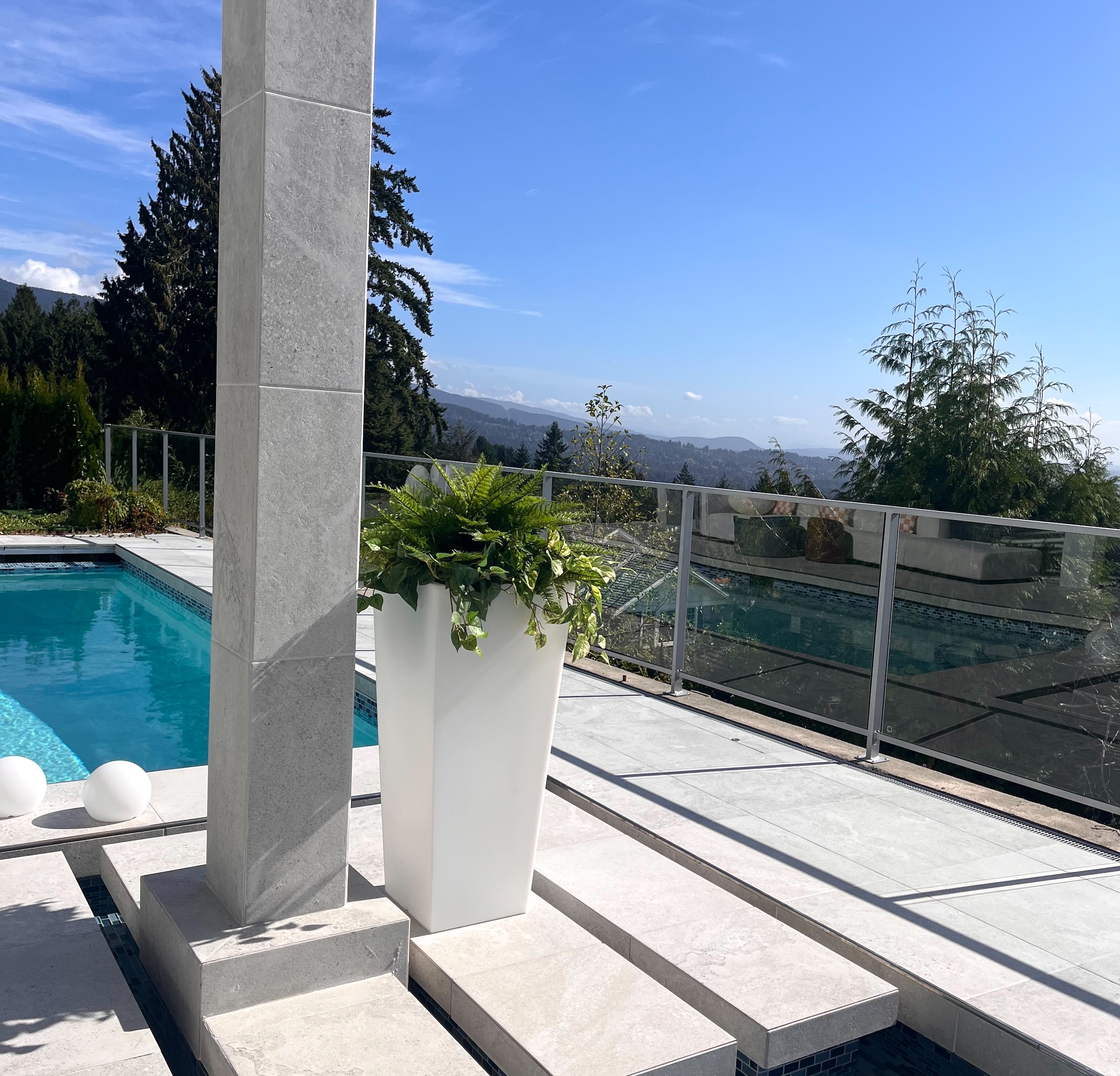 Tall white planter filled with artificial ferns and trailing faux greenery beside an outdoor pool and glass railing.