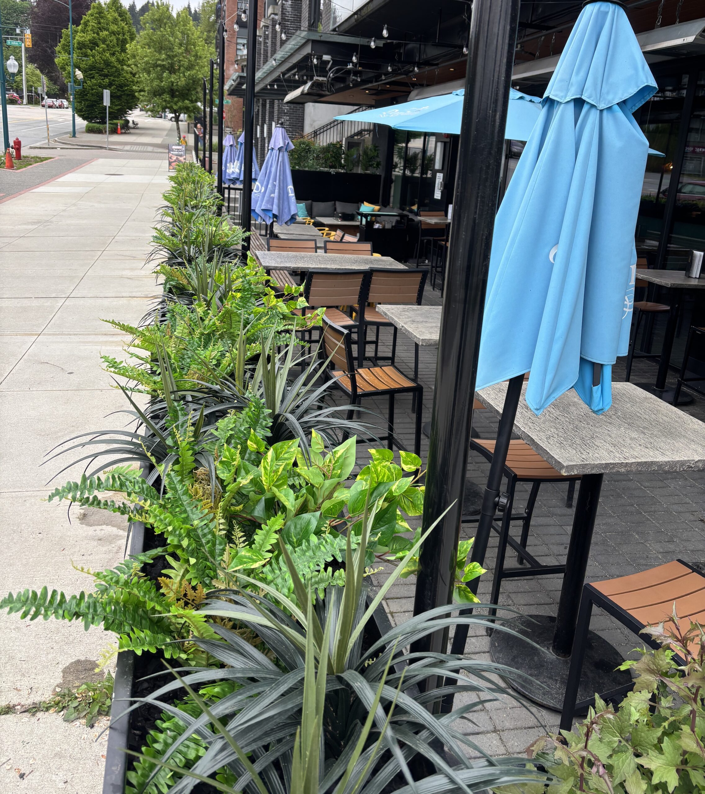 Long black planter boxes with artificial ferns, trailing faux foliage and spiky grasses line an outdoor restaurant patio beside tables and umbrellas.