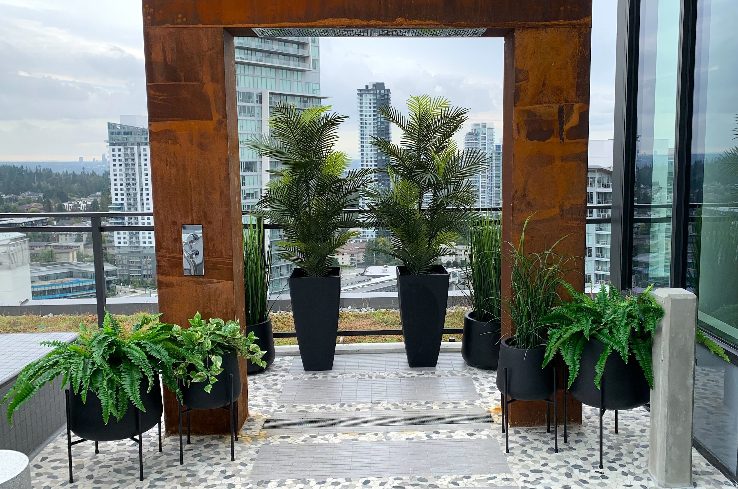 Black outdoor planters filled with artificial palms, grasses, ferns and trailing faux greenery on a rooftop pool deck.