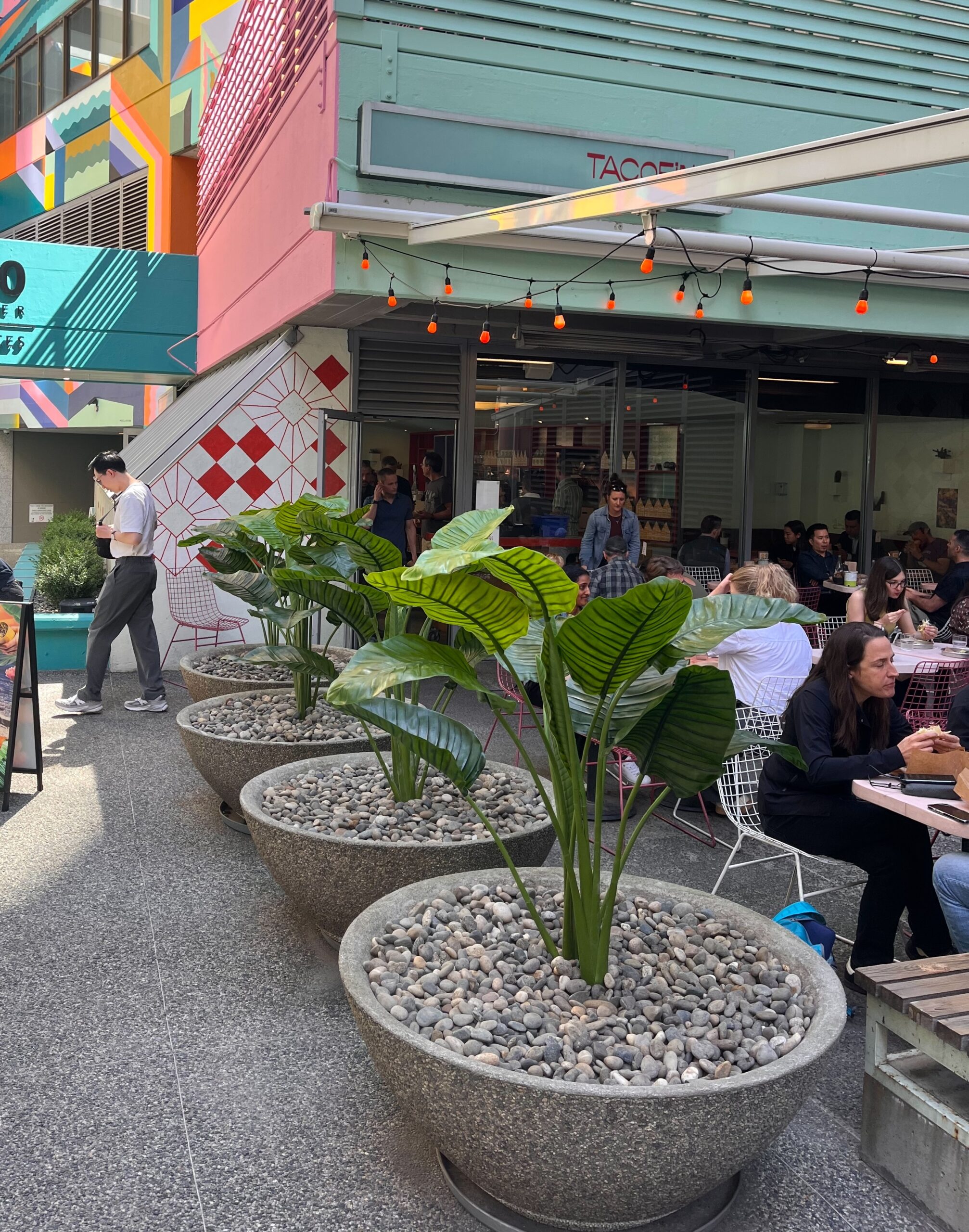 Large concrete planters filled with artificial tropical plants line an outdoor restaurant patio beside dining tables and storefront windows.