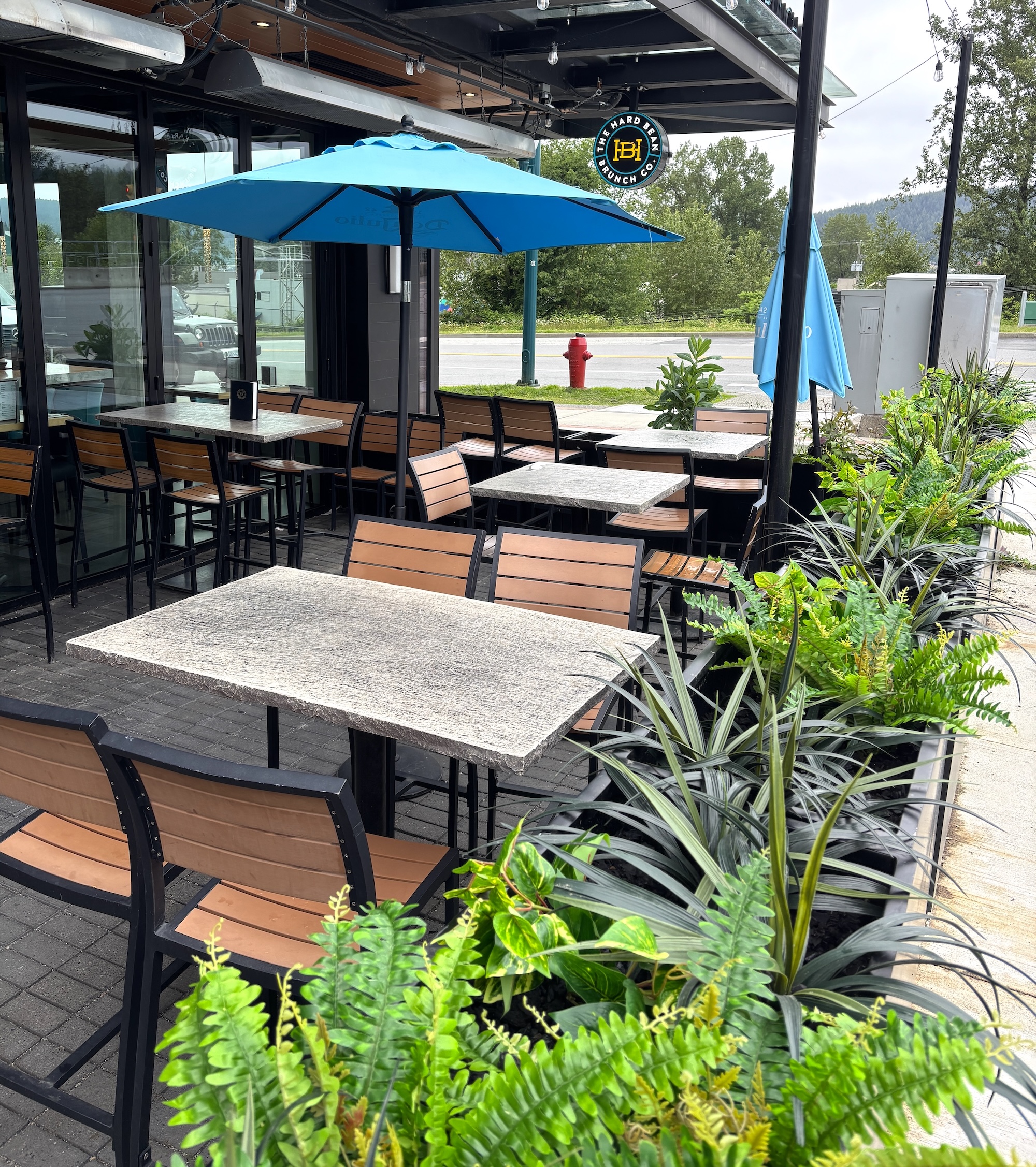 Black planter boxes filled with artificial ferns, spiky grasses and mixed faux foliage along an outdoor restaurant patio with tables and umbrellas.