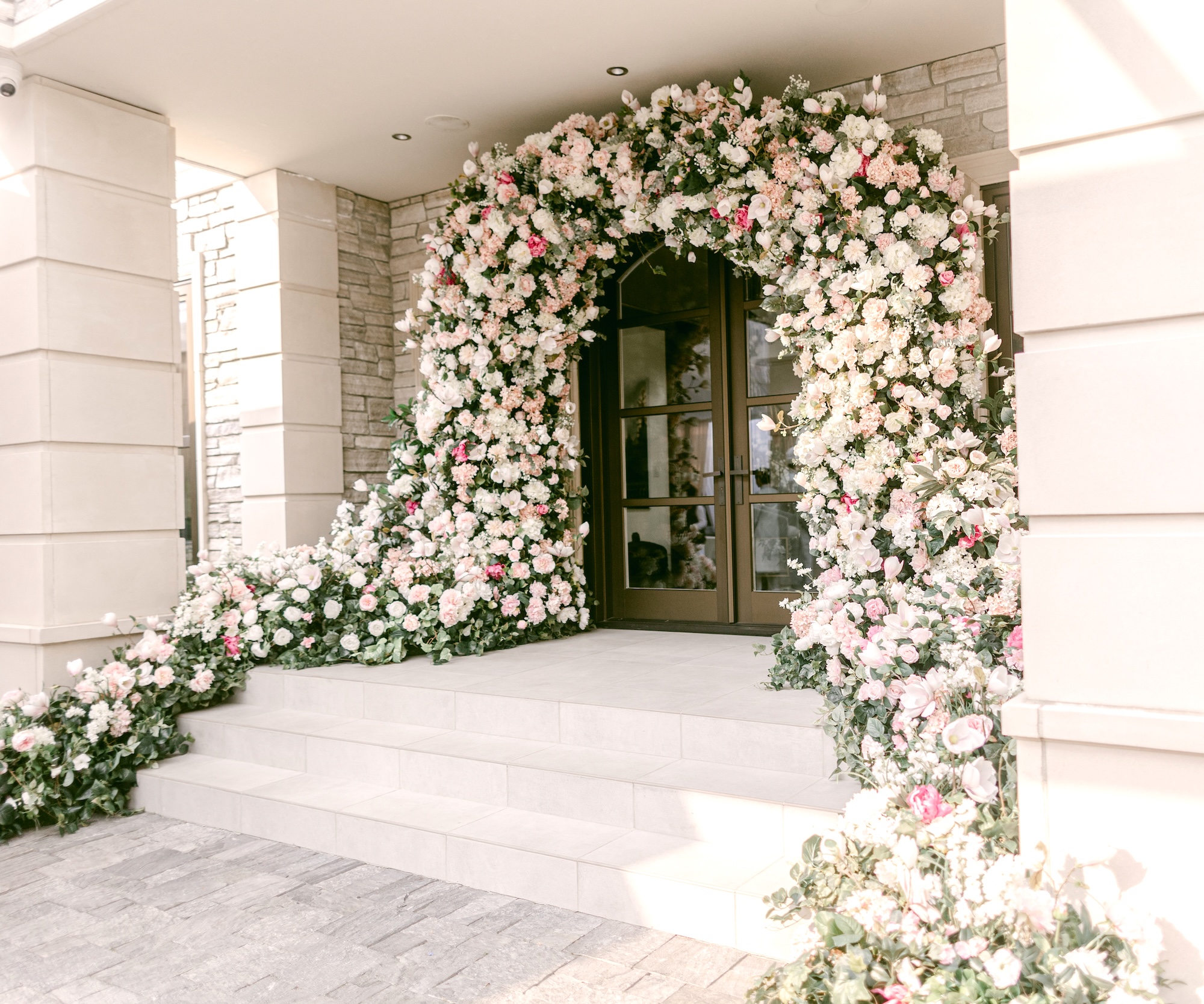 Large floral arch installation in white, blush, and soft pink flowers surrounding a stone venue entrance with cascading florals along the steps.