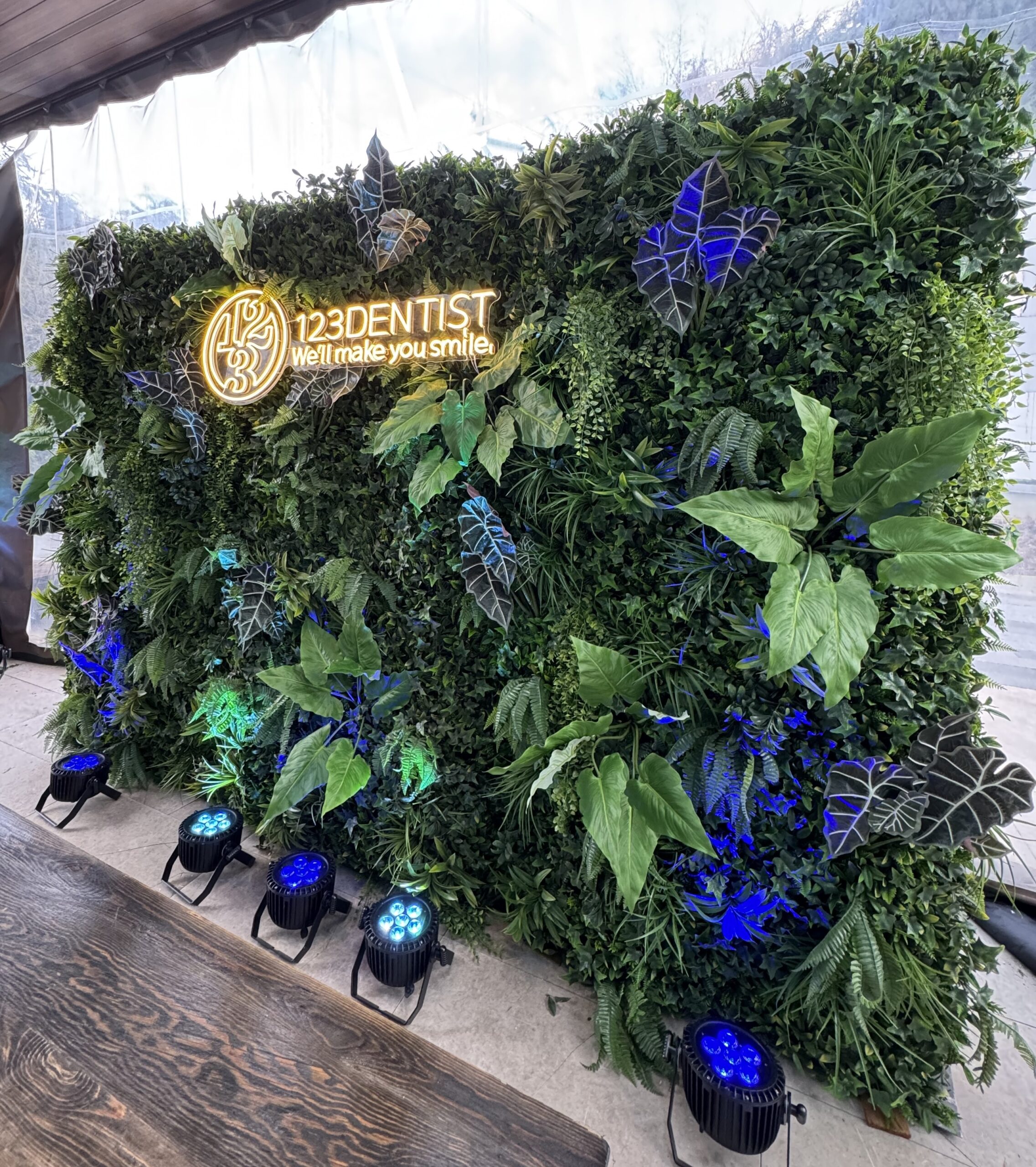 Tropical greenery wall with mixed faux leaves, oversized foliage, trailing plants, and an illuminated sign, lit with blue and green uplighting.