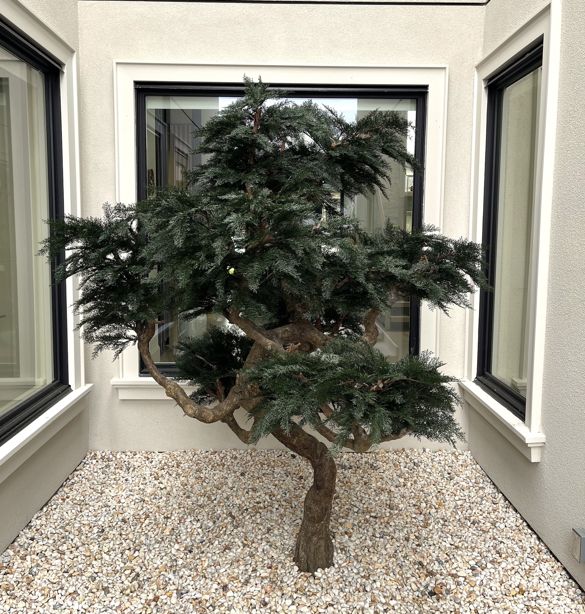 Artificial bonsai tree with a twisted natural-look trunk and dense green foliage set in a white gravel courtyard between windows.