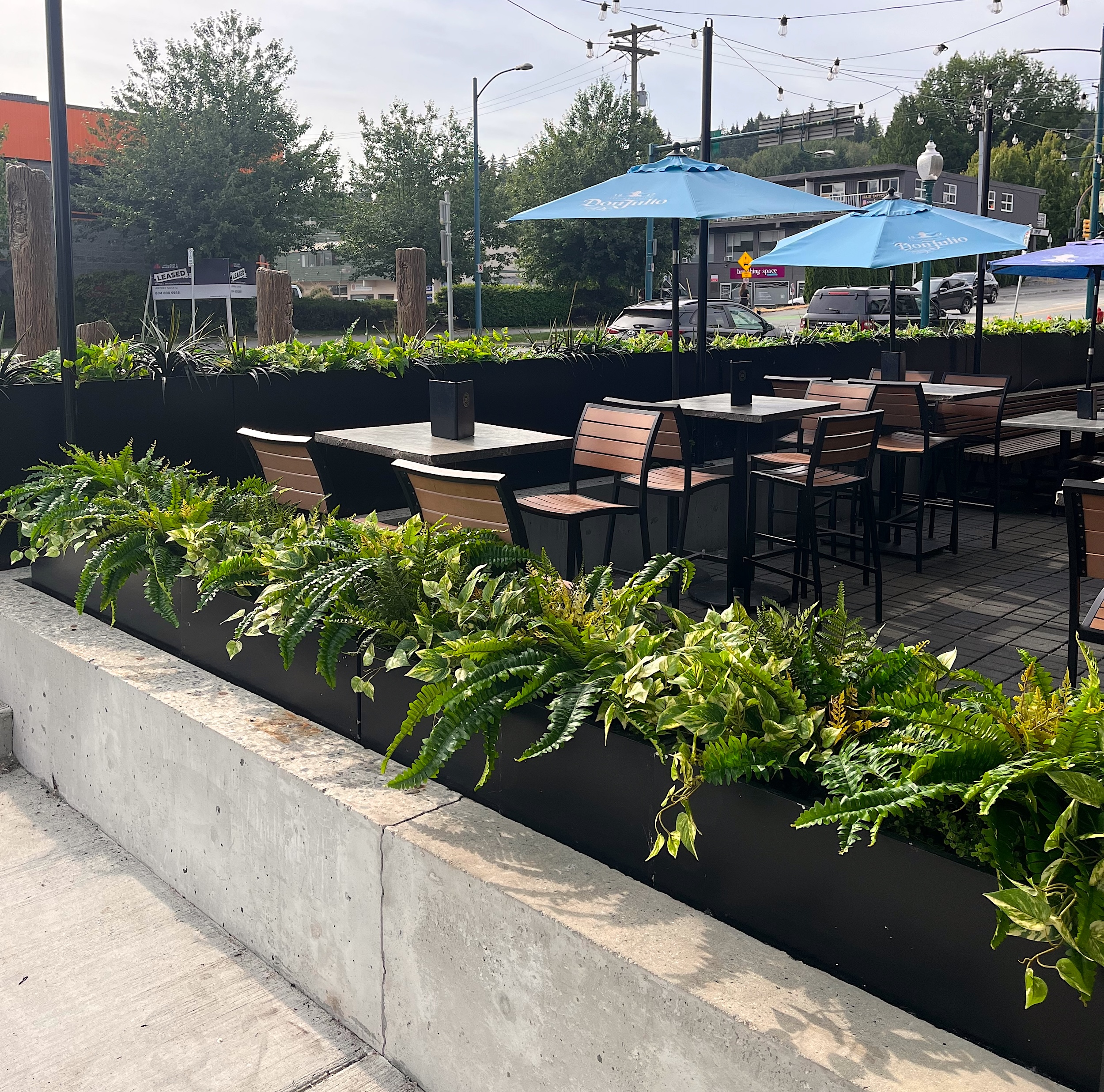 Black rectangular planter boxes filled with artificial ferns, variegated trailing foliage and spiky greenery border an outdoor restaurant patio.