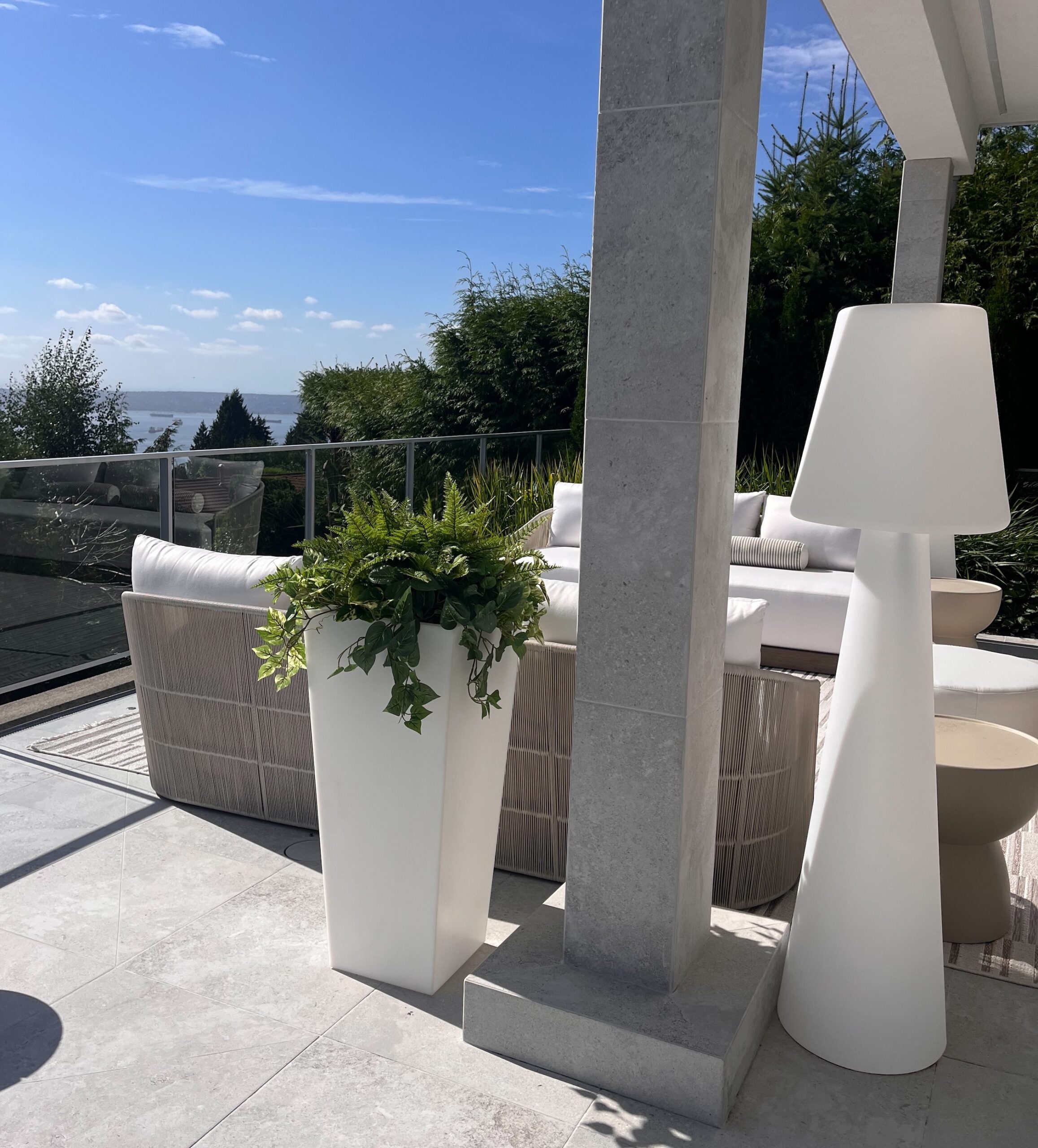 Tall white planter filled with faux ferns and trailing greenery on an outdoor pool deck beside white lounge seating.