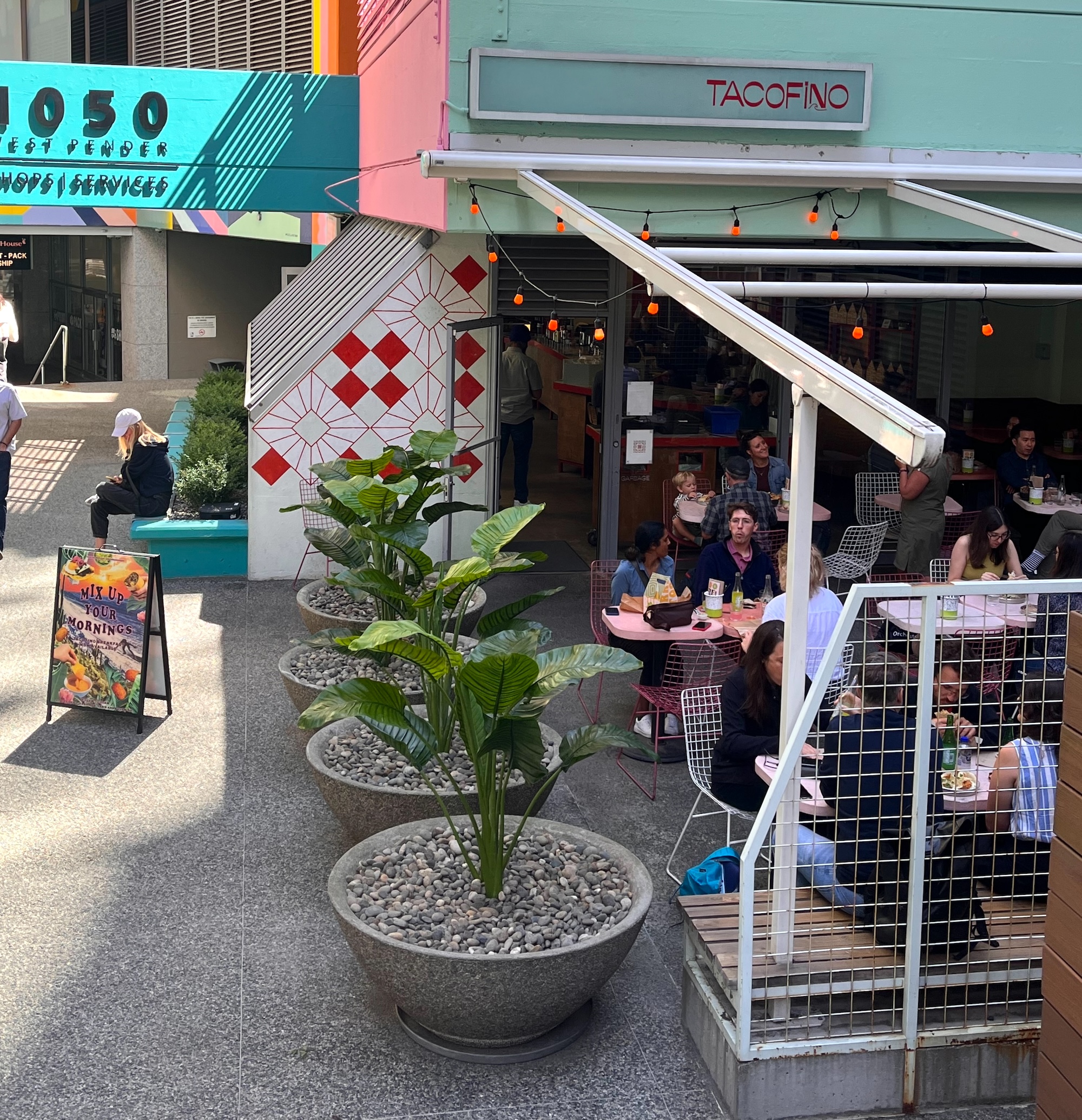 Large round planters with artificial tropical palms line an outdoor restaurant patio beside café tables and storefronts.