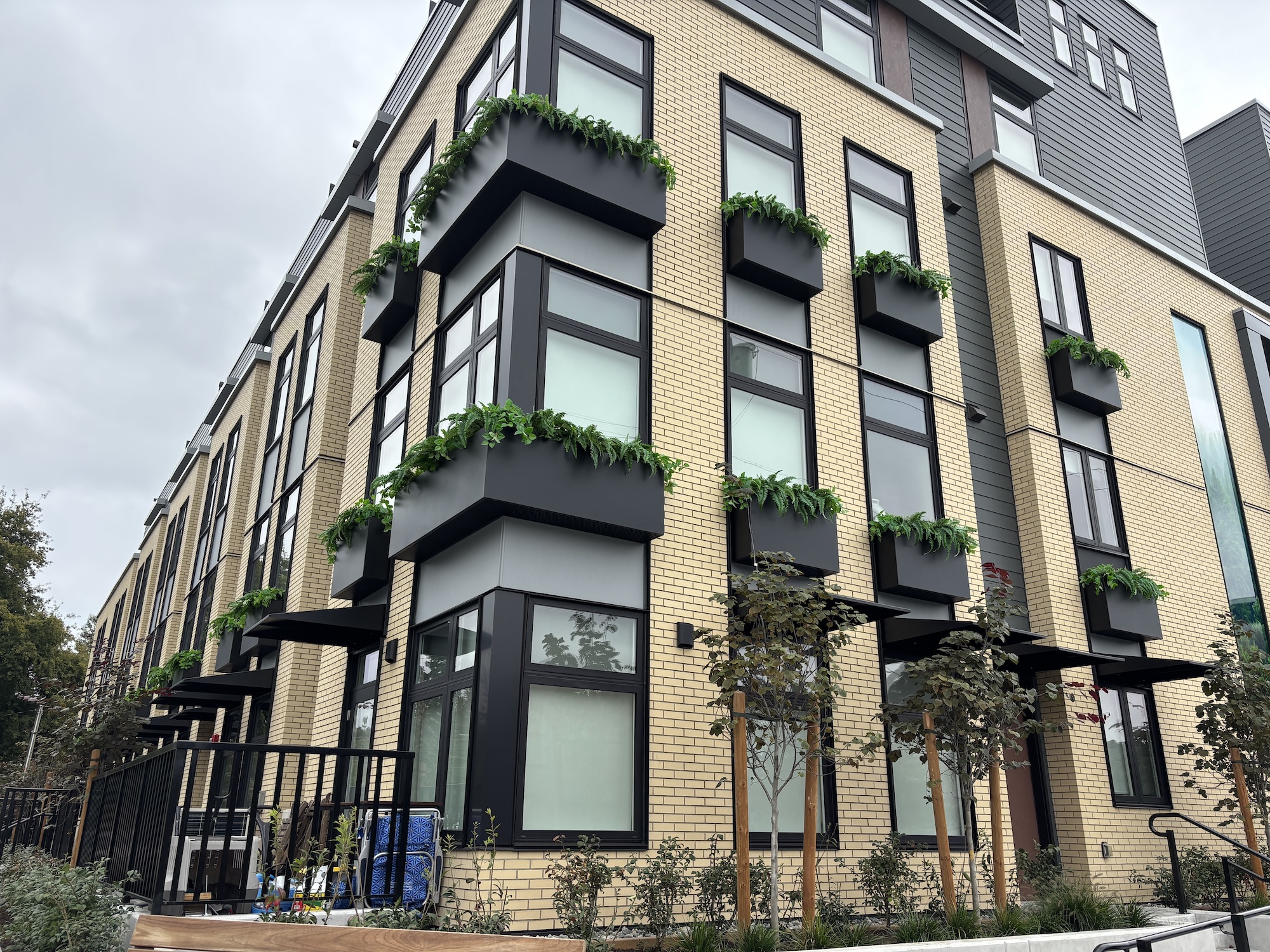 Black exterior window boxes filled with faux ferns and trailing greenery mounted across a multi-storey brick building façade.