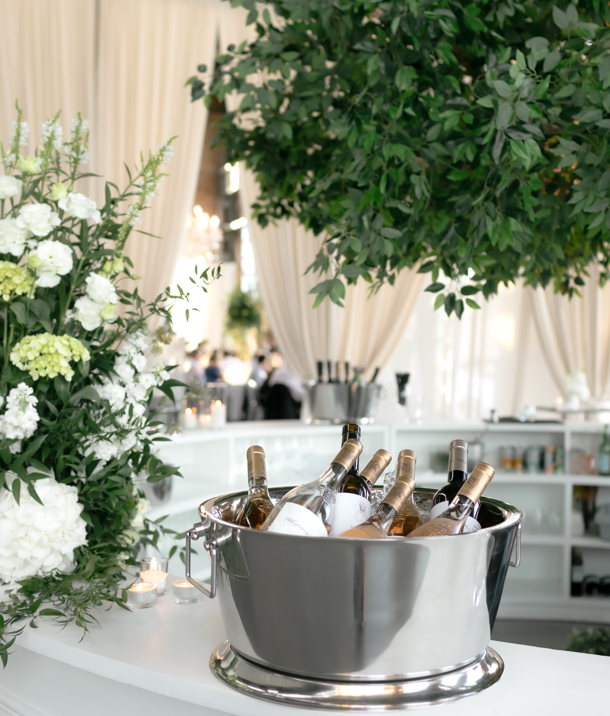 Large artificial tree canopy suspended above a white bar, with white floral arrangements, soft draping, and a silver wine bucket in an indoor event space.