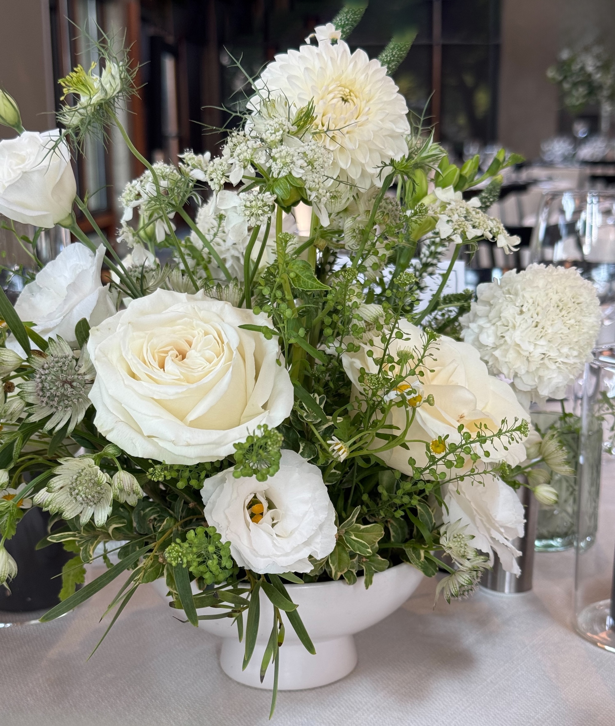 White floral centrepiece in a low white pedestal bowl with roses, lisianthus, dahlia, lace flower, and layered green stems on a reception table.
