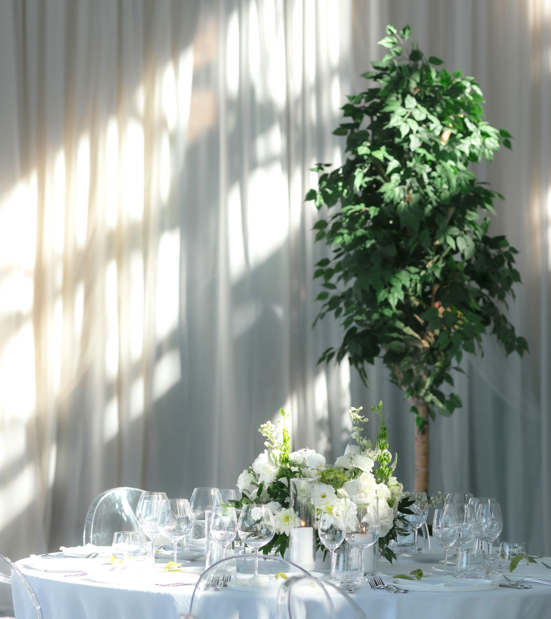Tall artificial leafy tree beside a round reception table with white floral arrangements, glassware, and soft white draping in an indoor venue.