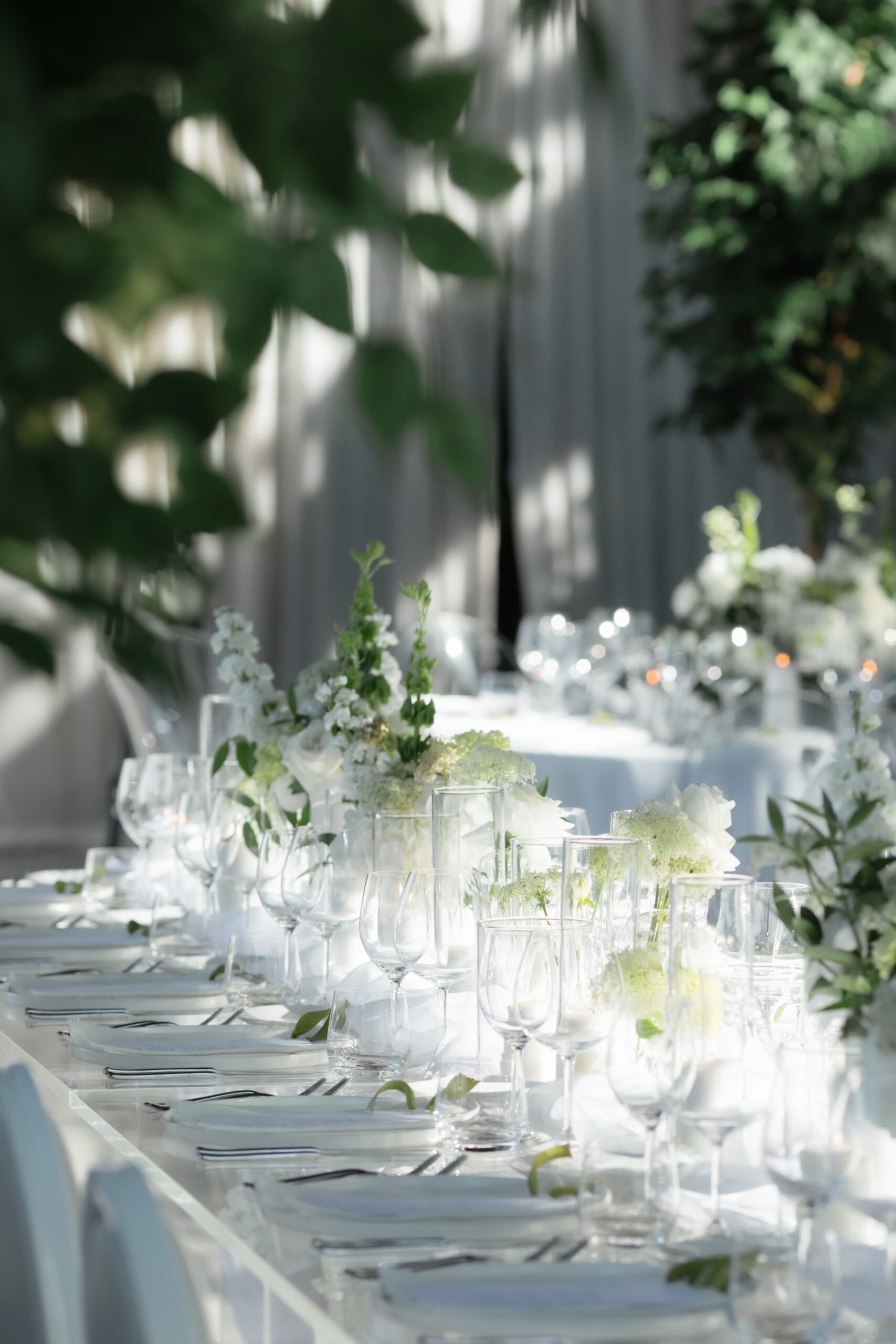 Long reception table with white floral arrangements in clear glass vases, layered place settings, and greenery accents in a softly lit indoor venue.