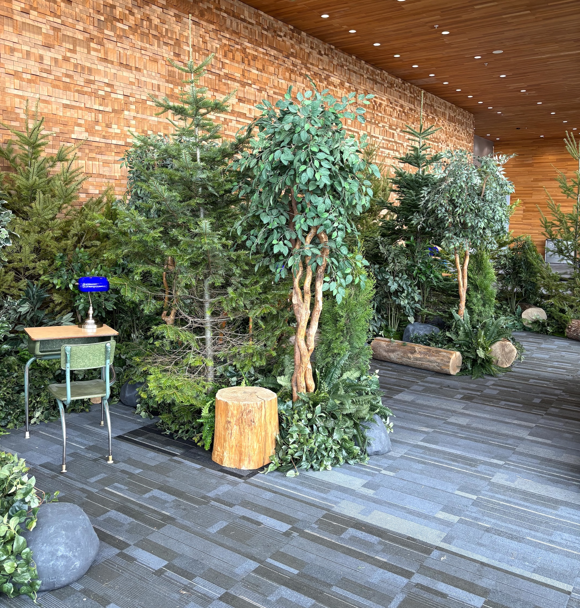 Forest-style backdrop with evergreen trees, leafy greenery, ferns, rocks, wood log accents, and a desk vignette in a corporate event venue.