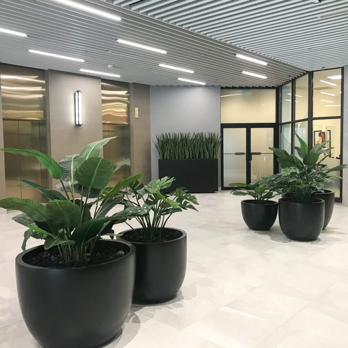 Large artificial tropical plants in black round planters arranged in a modern commercial lobby with marble flooring